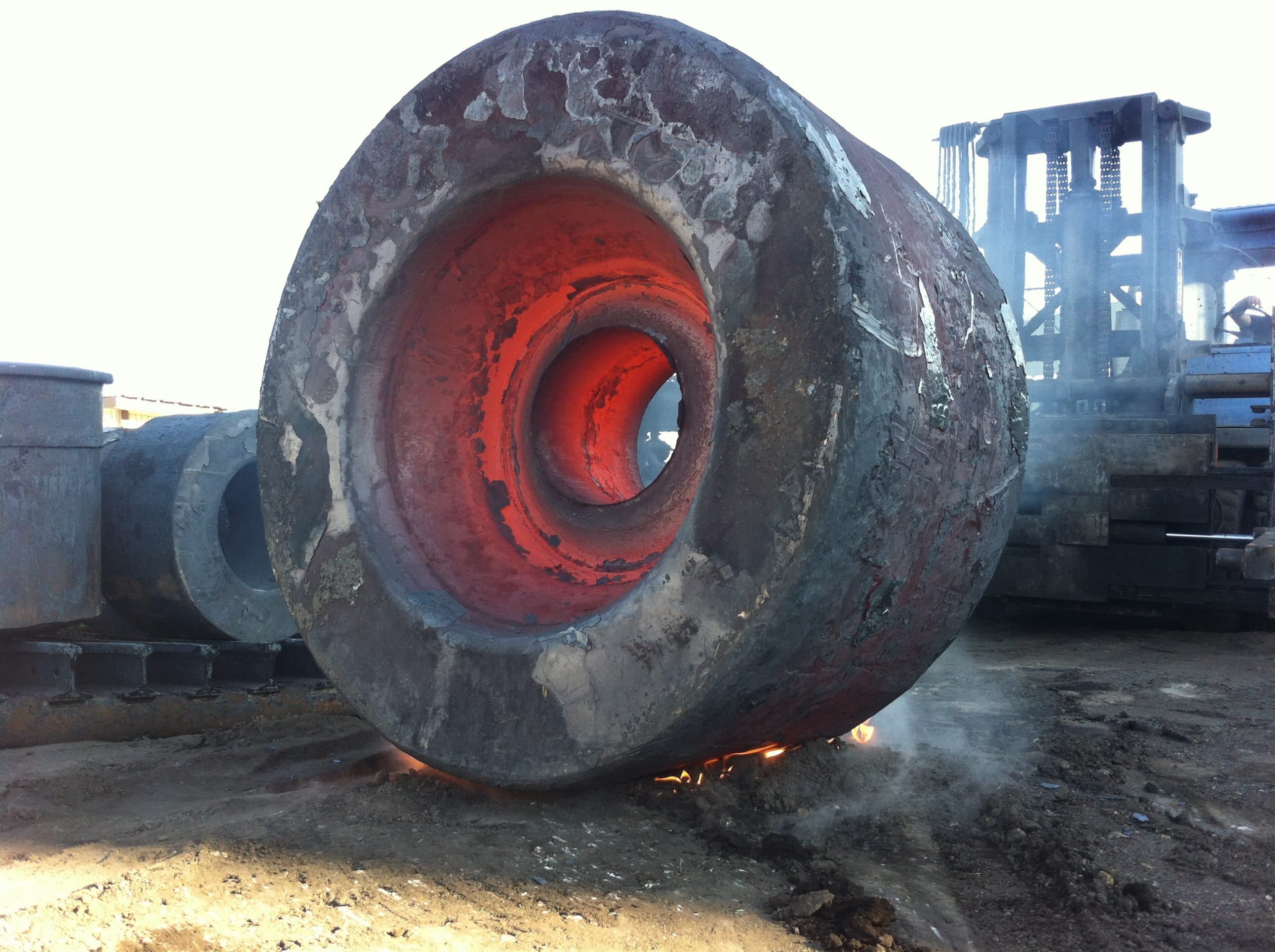 A large hollow forging with a glowing core cooling outdoors beside a manipulator